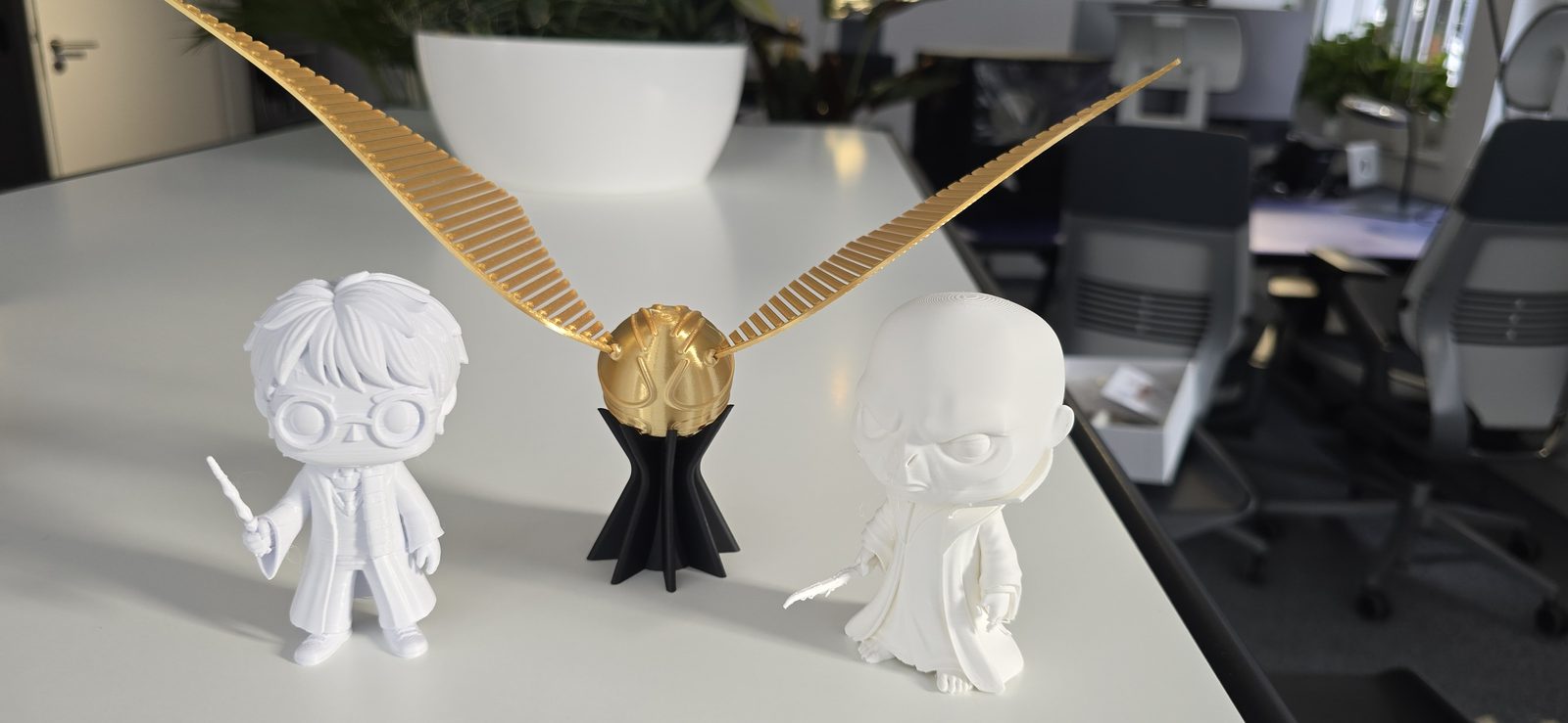 Drei weiß gedruckte Sammelfiguren und eine goldene, geflügelte Kugel auf schwarzem Standfuß