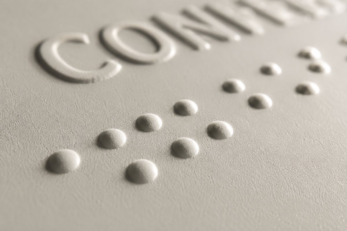 Nahaufnahme eines hellen Schildes mit erhabener Großbuchstaben-Schrift und daneben deutlich gewölbten Braille-Punkten, beide im Relief aufgedruckt